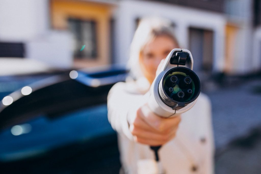 Imagem desfocada de uma mulher segurando um carregador de carro elétrico. O centro do carregador está focado, onde identificamos sua forma circular, com 5 pinos a vista, também circulares, sendo 3 mais grossos e 2 mais finos.