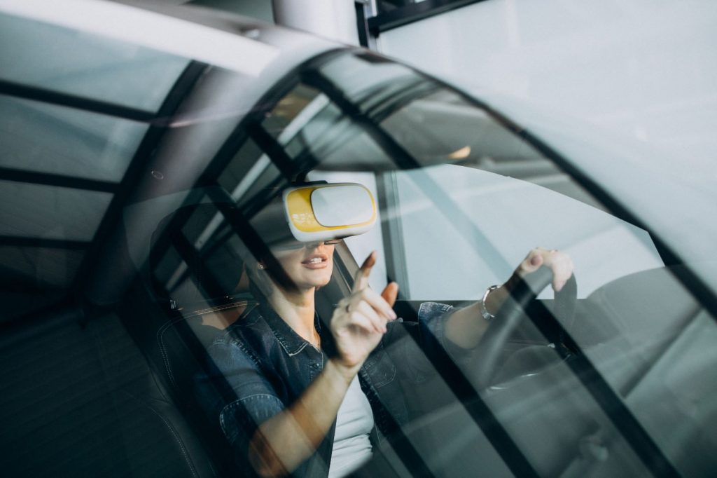 Imagem de uma mulher dentro de um carro, vista através do vidro do painela. Ela está com uma mão segurando no volante e a outra apontando para o vidro.
