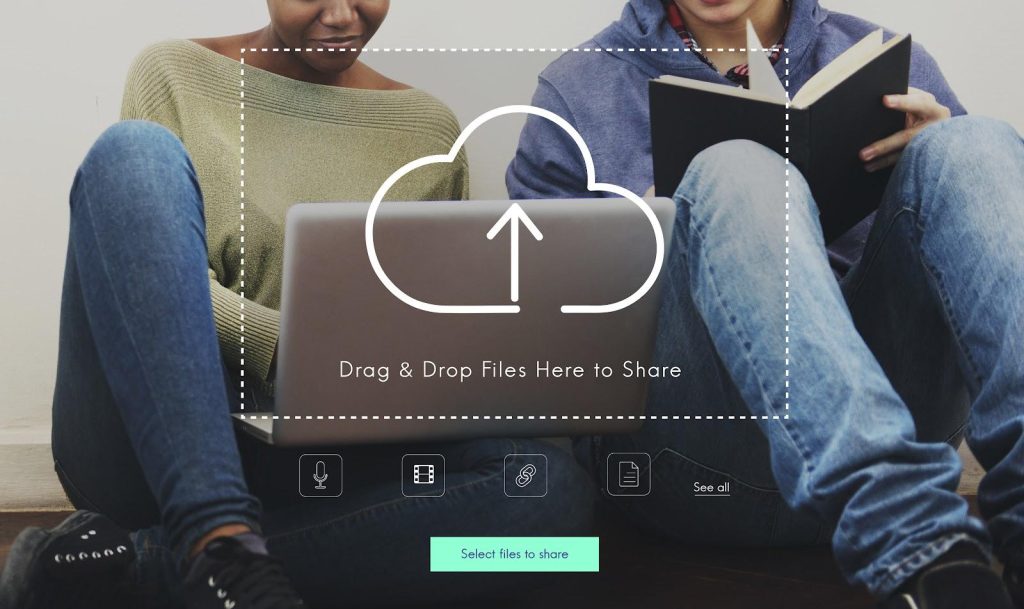 um homem e uma mulher estão sentados no chão de um local, encostados em uma parede branca e com um notebook no colo. Ambos olham para a tela do notebook; A mulher tem as duas mãos no teclado e o homem segura um caderno preto. Sobrepondo a imagem, está um símbolo de upload com a frase “Drag & Drop Files Here to Share”.