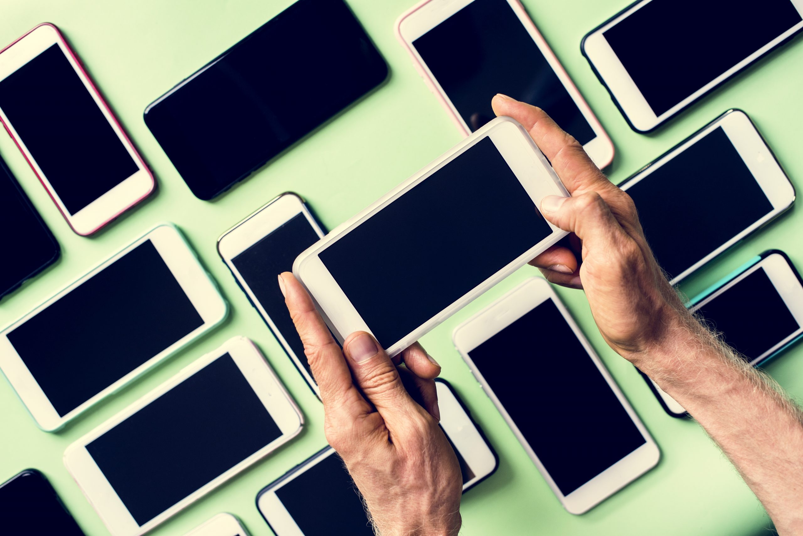 Foto de diversos smartphones sobre uma superfície verde. Acima deles, vemos um par de mãos segurando um destes dispositivos. A imagem tem como objetivo ressaltar a importância das boas práticas de descarte de eletrônicos