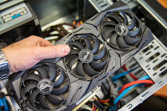  Uma mão segurando uma placa de vídeo com três ventoinhas, enquanto é instalada em um computador. Fios e componentes internos são visíveis ao fundo.