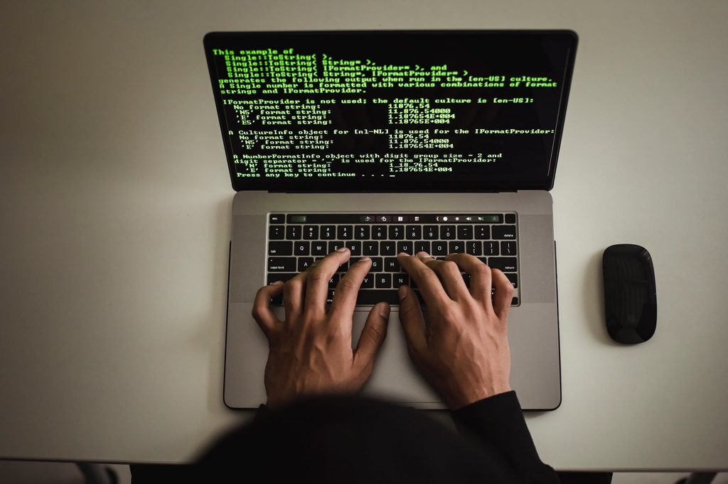 Notebook aberto em cima de mesa branca com tela ligada em códigos de programação. Pessoa de blusa preta digitando no notebook. 