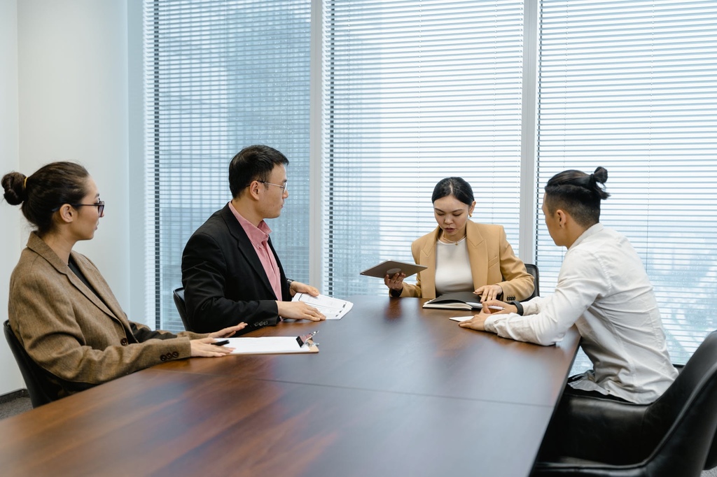 Grupo de quatro pessoas, sendo dois homens e duas mulheres, sentados ao redor de uma mesa de trabalho, conversando e manuseando papéis.