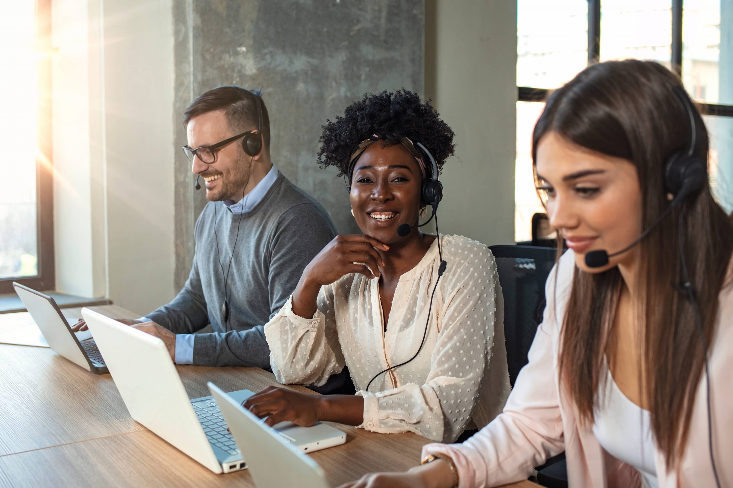 três pessoas de frente para um computador e com fones aos ouvidos. Ao centro, uma mulher negra olha para o centro da imagem e sorri