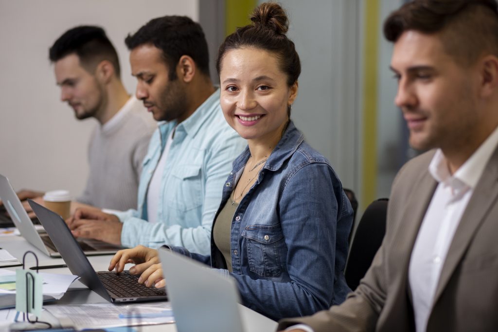 Profissional da tecnologia olhando para a câmera enquanto sorri e utiliza notebook. Ao seu lado, outros profissionais de TI utilizam notebooks.