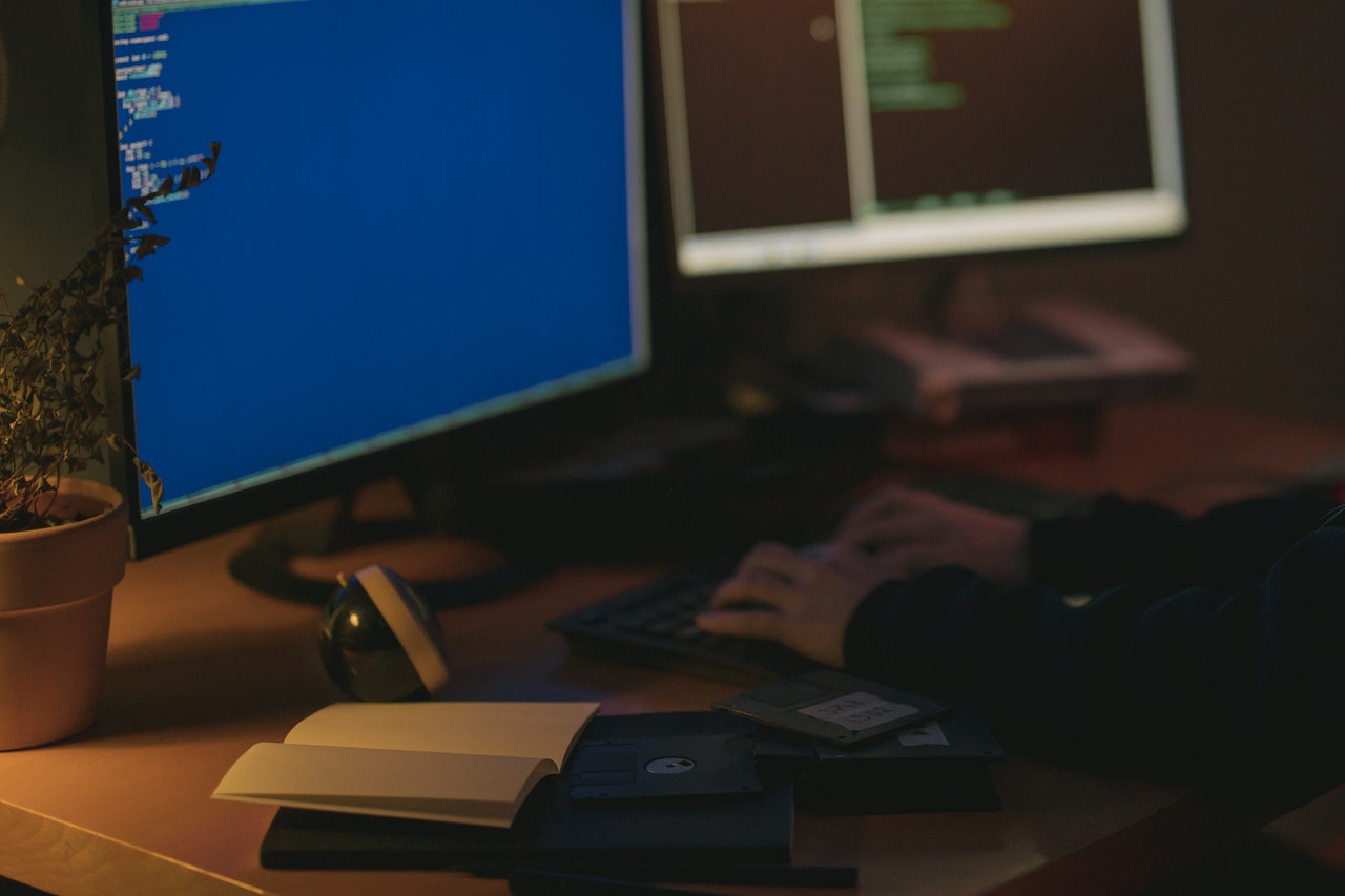 Pessoa de blusa preta digitando no teclado de um computador que está sobre mesa de madeira. Em cima da mesa tem-se também livros, disquetes e uma planta.