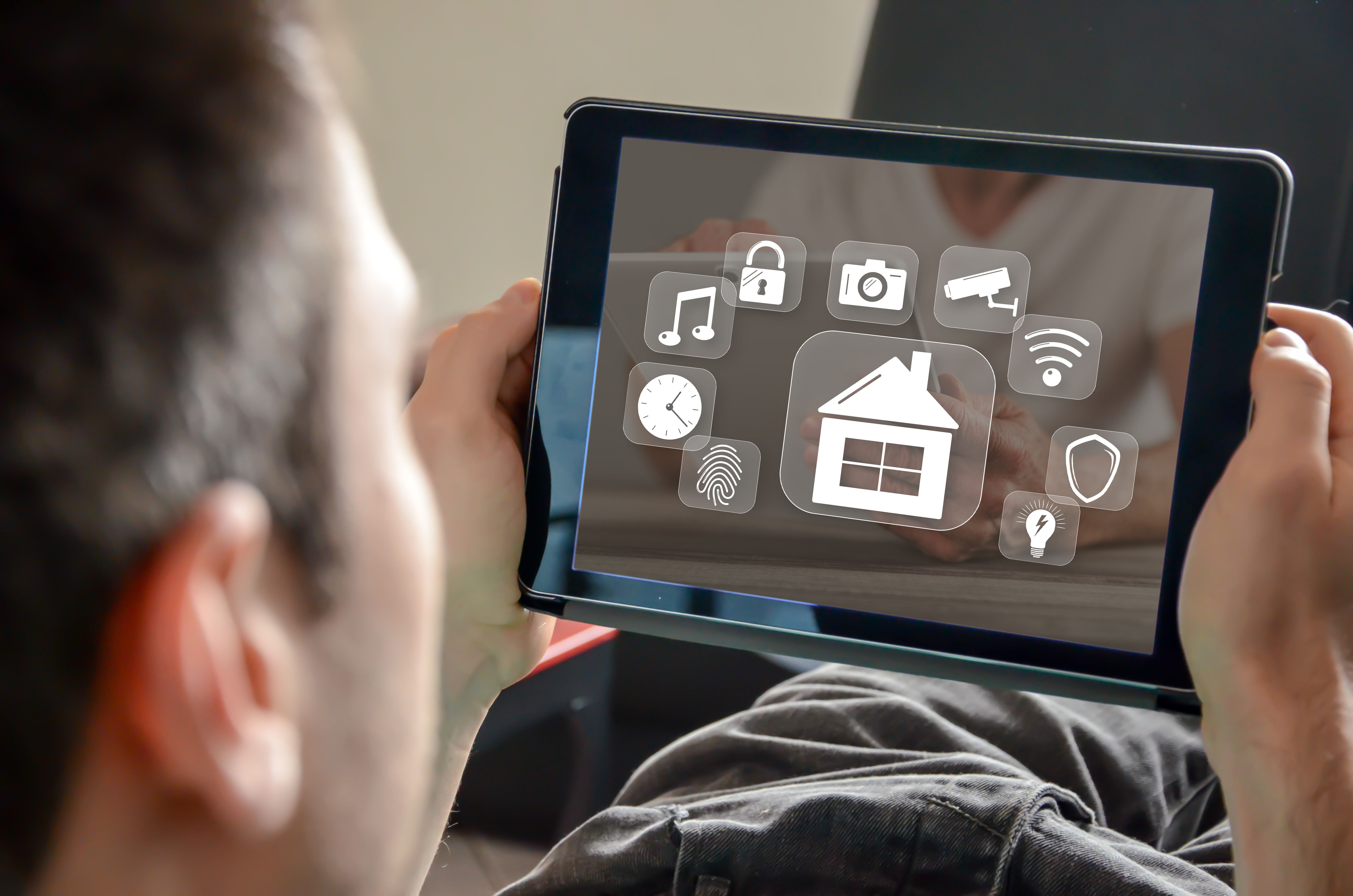 Imagem com um home deitado e de costas segurando um tablet, na tela há o desenho de vários símbolos como o de uma casa, uma lâmpada, uma digital, um relógio, uma nota musical, um cadeado, uma câmera fotográfica, uma câmera de segurança, um wi-fi e um escudo. Todos estão simbolizando uma smart home.