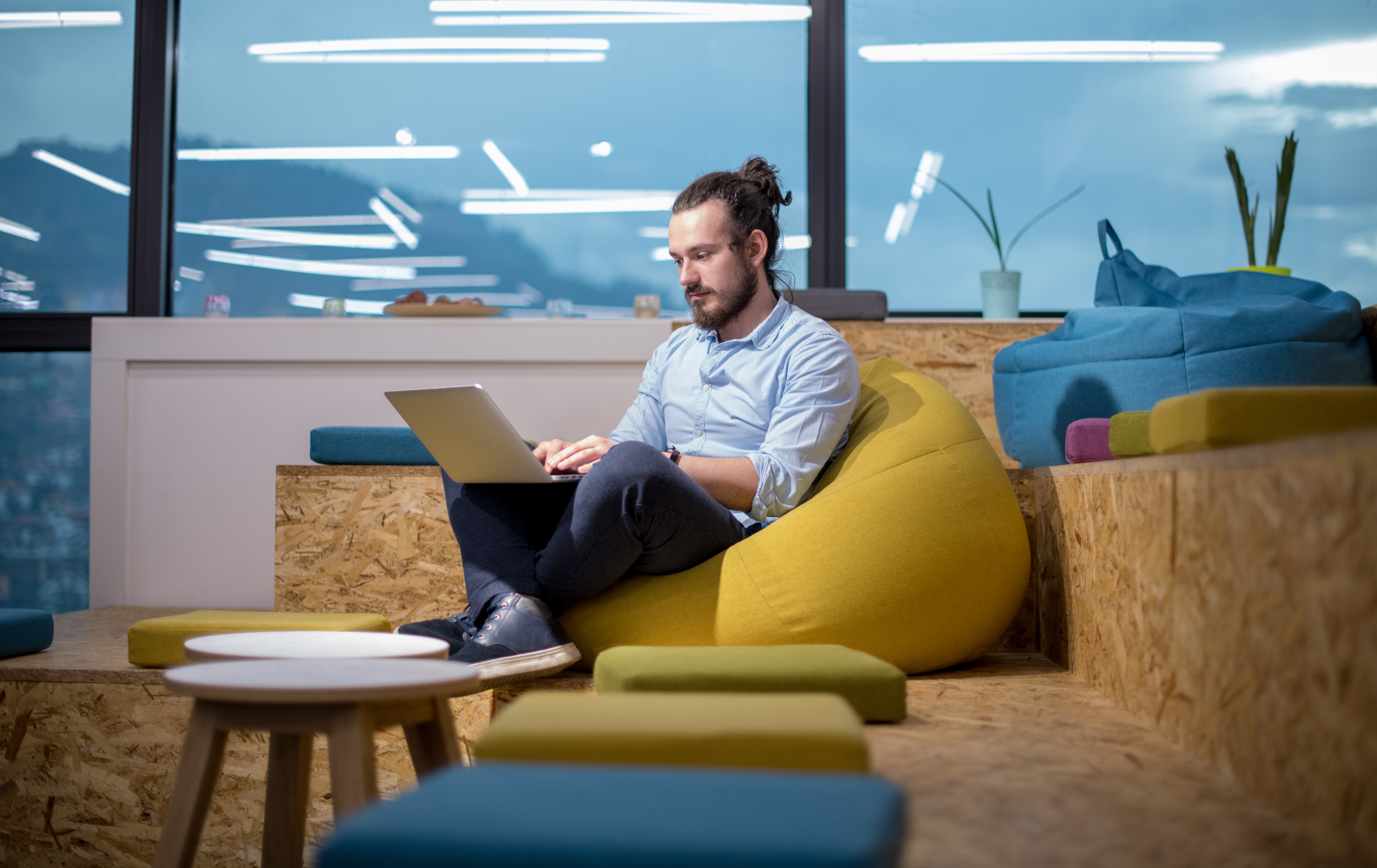 Imagem de uma sala de uma empresa com paredes transparentes que permitem a vista do lado de fora, banquinhos coloridos (verde, amarelo e azul) espalhados pelos espaço, um puff amarelo e outras almofadas espalhadas, no centro há um homem sentado no puff e no colo dele há um computador. 