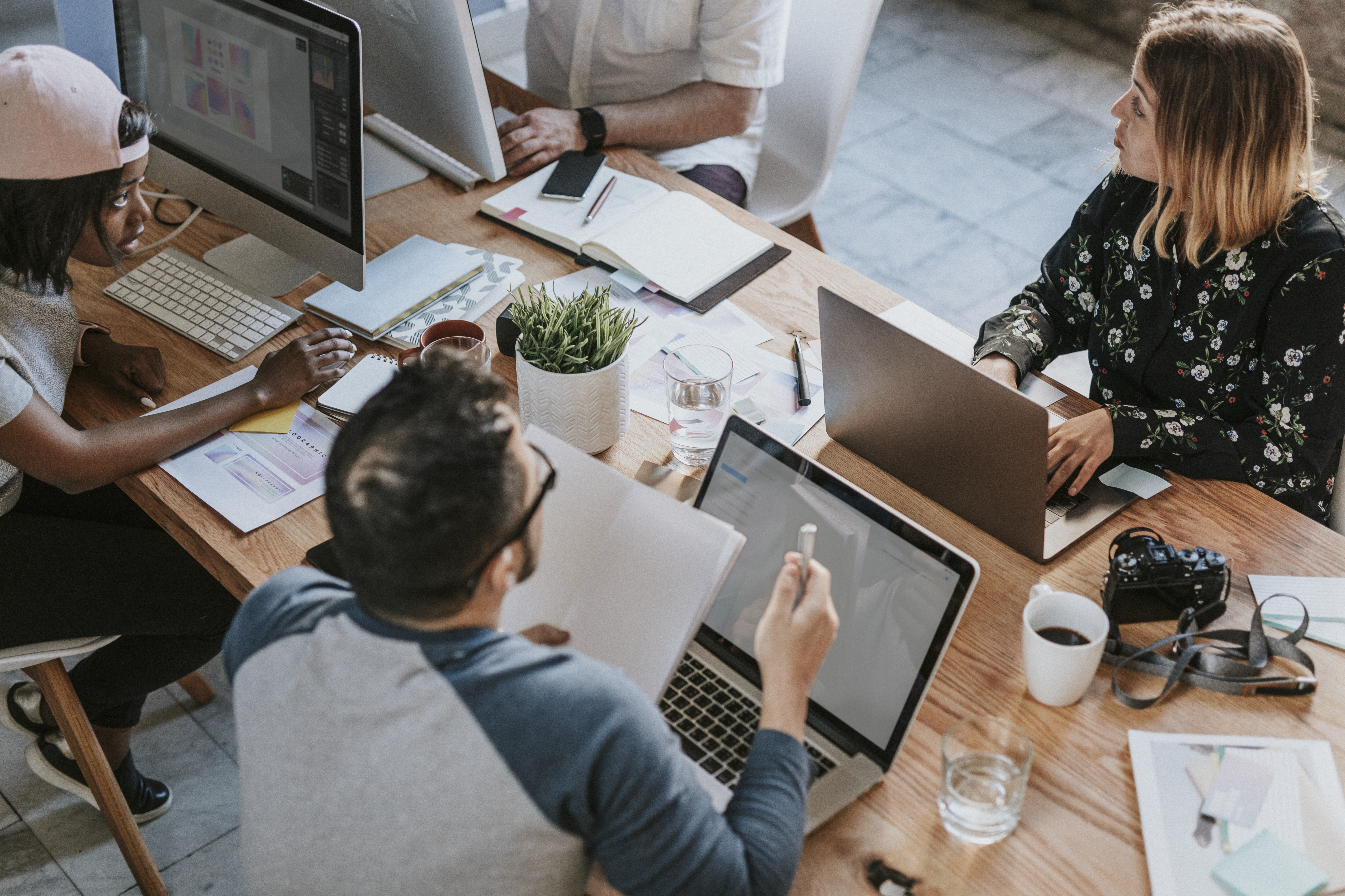  Imagem de uma sala de trabalho, nela há uma mesa de madeira clara com uma equipe de quatro pessoas trabalhando em computadores, em cima do móvel há também xícaras de café, copos de água, cadernos, folhas, agendas, canetas, uma câmera fotográfica e um vaso com uma planta.