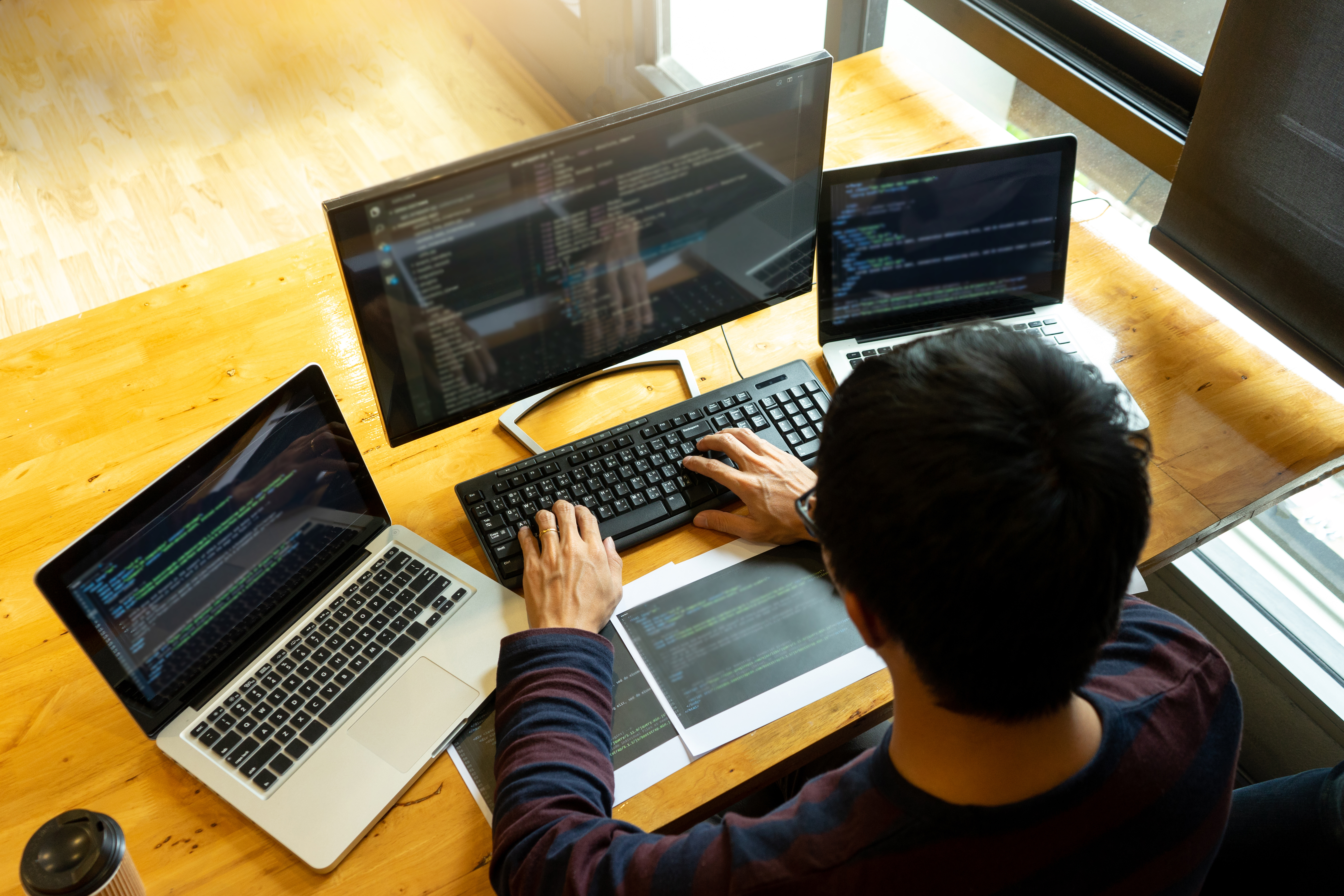 Da esquerda para a direita: um copo de café em cima de uma mesa de madeira clara, em cima da mesma há um notebook com tela preta e códigos coloridos nela, depois há um teclado e um monitor também com códigos coloridos e um homem sentado de frente e teclando, na frente dele há duas folhas impressas com códigos e no final há mais um notebook muito parecido com o primeiro. A imagem representa o trabalho de um desenvolvedor de software independente.