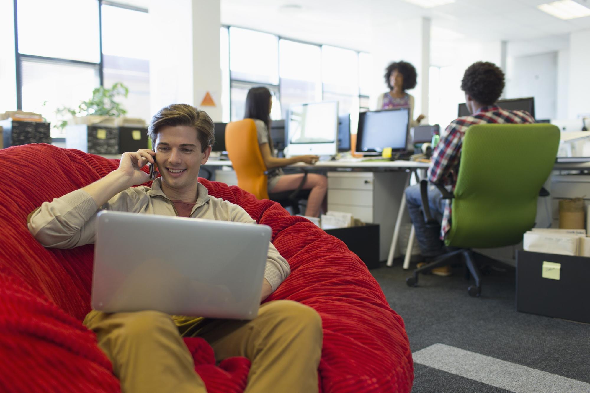 Imagem ambientada em um escritório de trabalho, do lado esquerdo há um pufe vermelho grande e em cima dele há um homem branco sentado, com um notebook no colo e com um celular na orelha sorrindo, do lado direito há três pessoas, duas sentadas em cadeiras de frente para os computadores e uma de pé, parecem estar conversando.