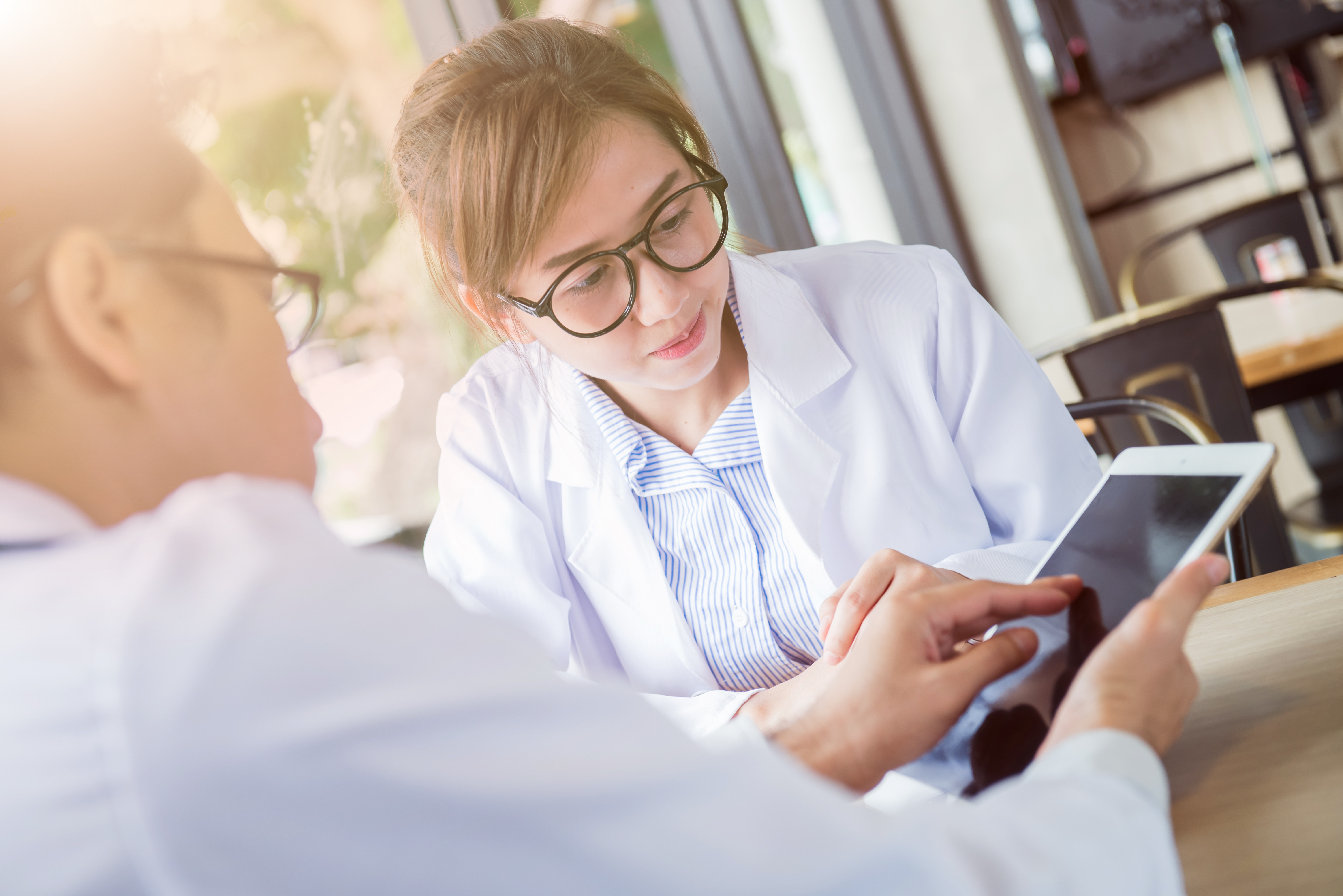 Do lado esquerdo há um homem vestindo roupas brancas, usando óculos e de costas para a câmera, do lado direito há uma mulher asiática de cabelos presos, usando óculos e olhando para o tablet que o homem está segurando e teclando
