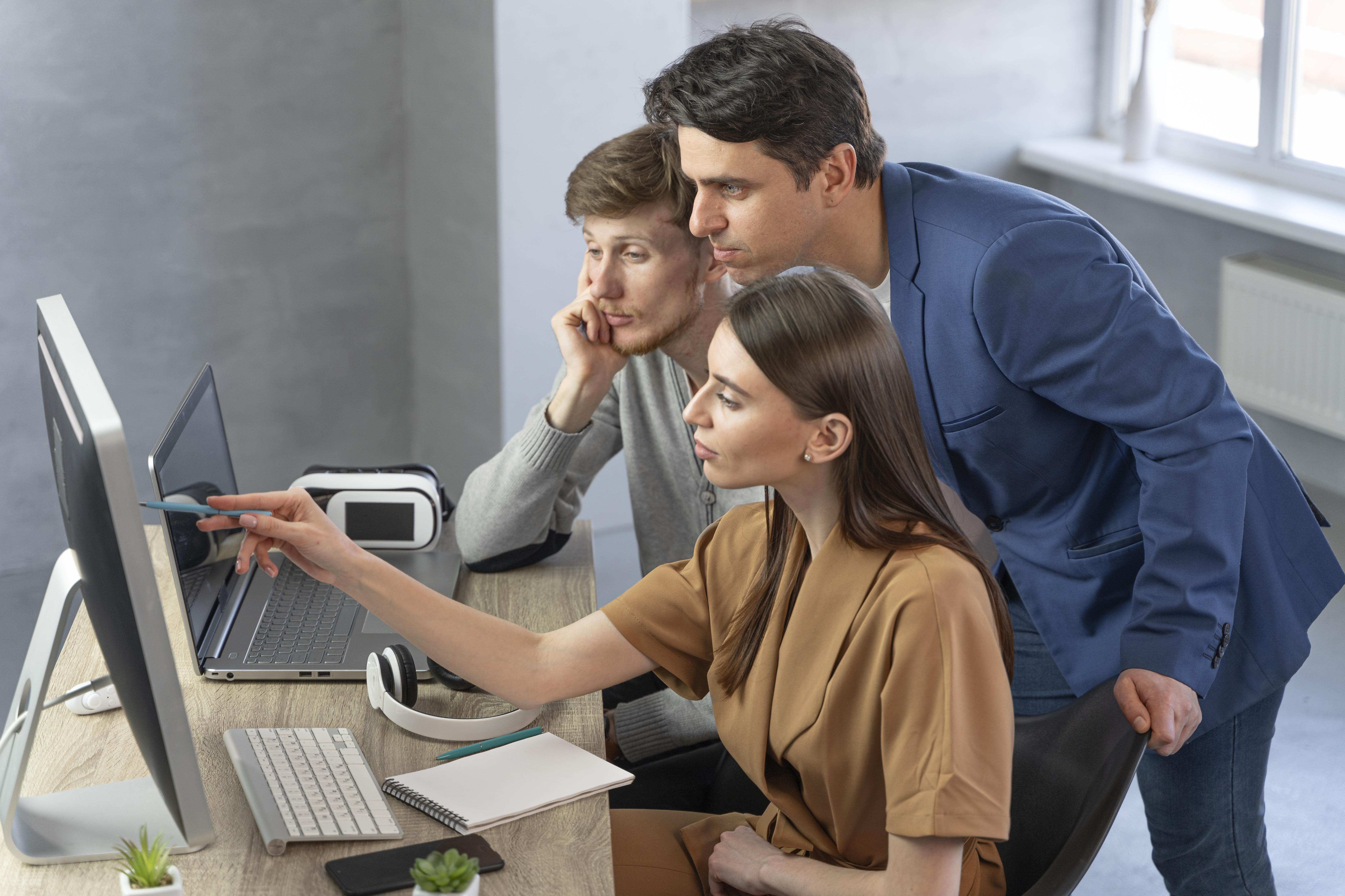 Um grupo de três pessoas, uma mulher sentada, um homem sentado e um homem de pé atrás deles estão olhando para a tela de um computador, em cima da mesa há um teclado, um fone de ouvido, um celular, caneta, caderno e um estojo. 