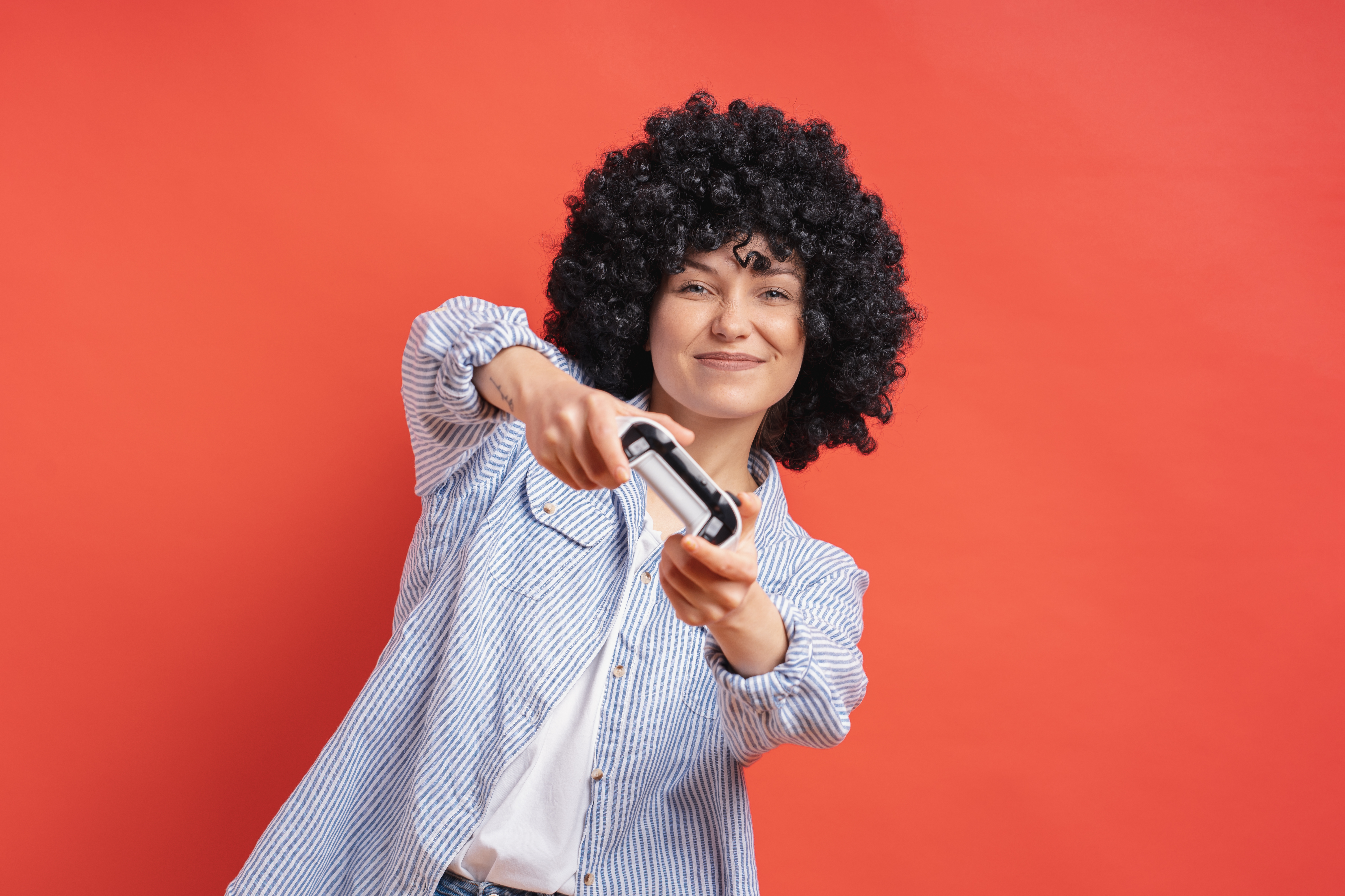 Imagem com uma parede vermelha ao fundo e, ao centro, há uma mulher de cabelos enrolados e se inclinando, ela está segurando um controle de console e está fazendo uma expressão de felicidade.