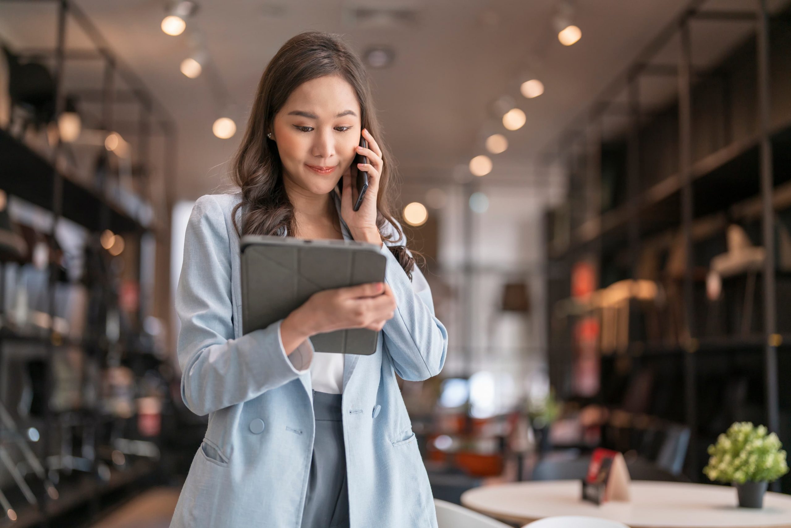 Mulher asiática com celular próximo da orelha olha para um tablet em suas mãos