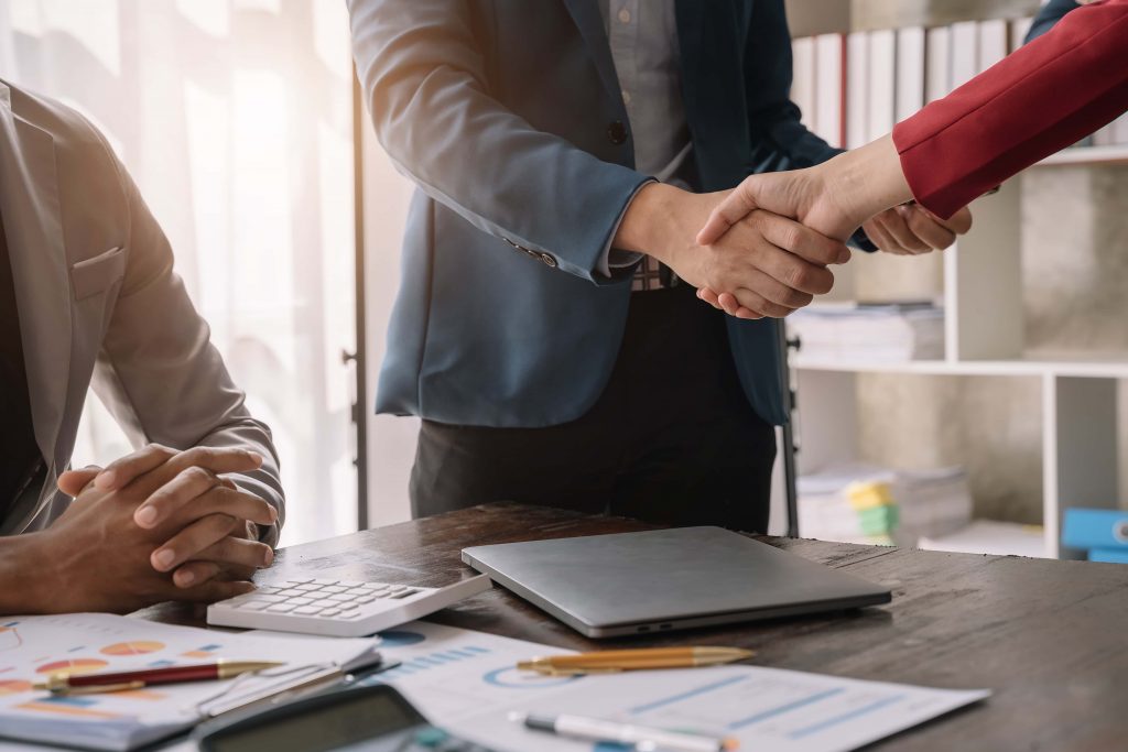 Mãos de duas pessoas brancas em cumprimento dentro de um escritório, com notebook e papéis sobre a mesa. 