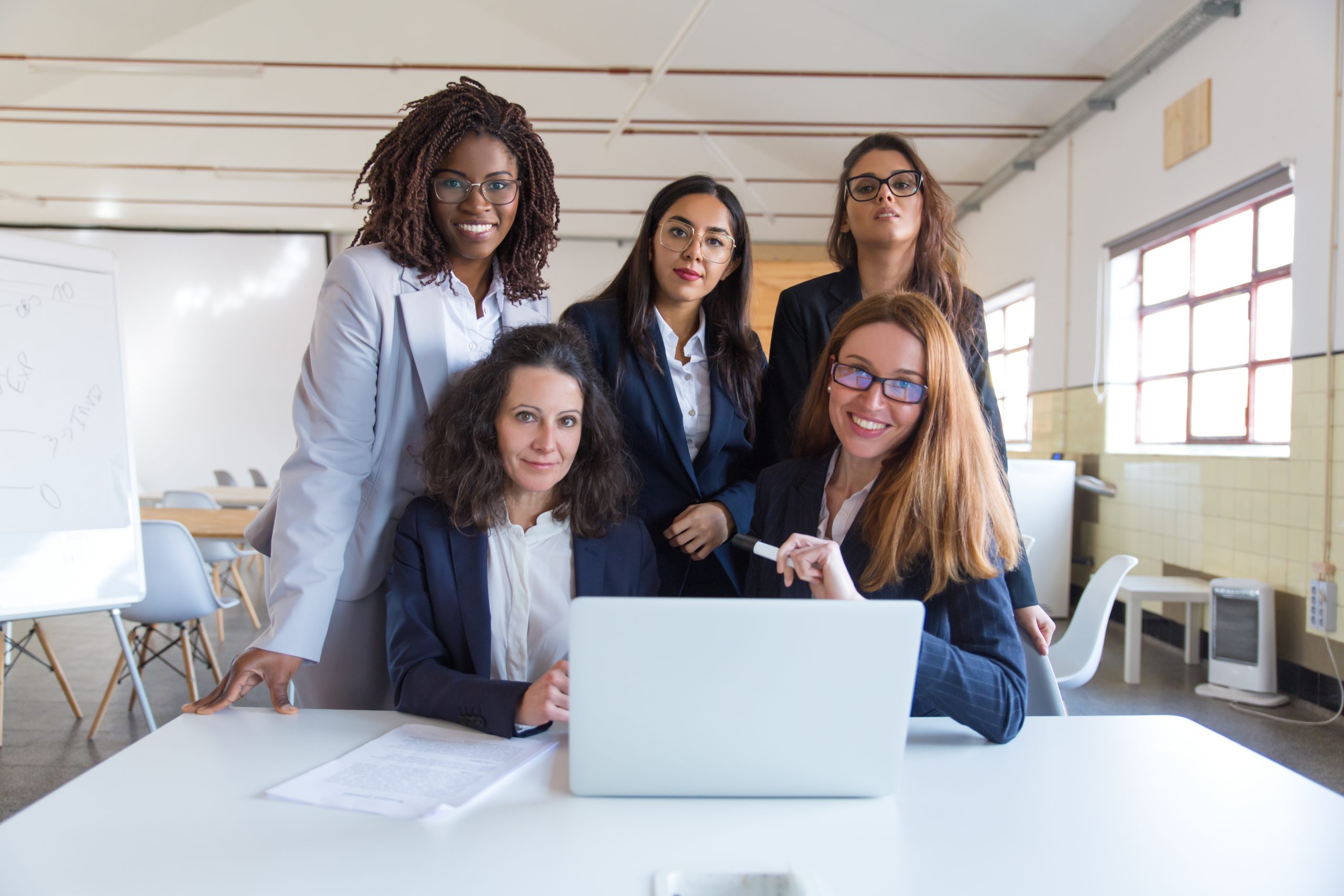 Imagem de uma fotografia em que há uma mesa branca com um notebook da mesma cor em uma sala, na frente do computador há duas mulheres brancas sentadas olhando para a câmera sorrindo e, atrás delas, há outras três mulheres. Representando as mulheres na tecnologia.