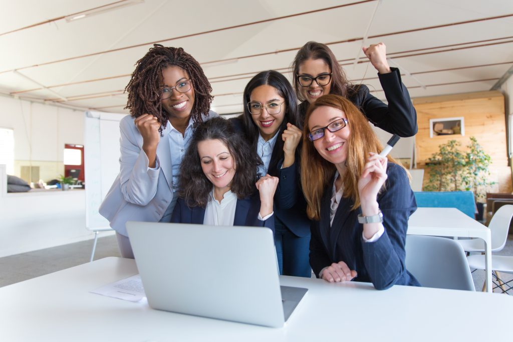 Imagem de uma fotografia em que há uma mesa branca com um notebook da mesma cor em uma sala, na frente do computador há duas mulheres brancas sentadas olhando para a câmera sorrindo e, atrás delas, há outras três mulheres, elas estão comemorando algo. A imagem está representando as mulheres na tecnologia.