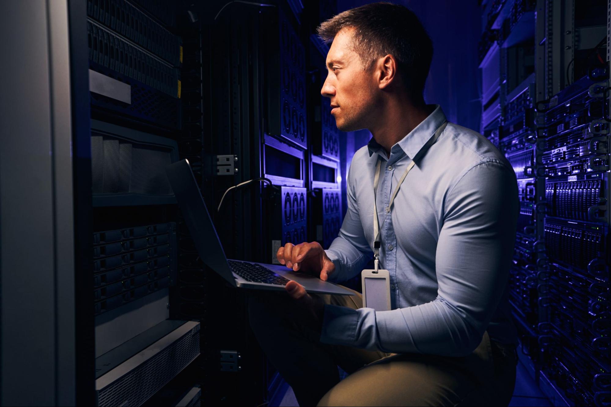 Um homem que veste camisa social azul e um crachá está agachado em um data center enquanto segura um notebook em suas mãos. Ele olha para a estrutura do data center
