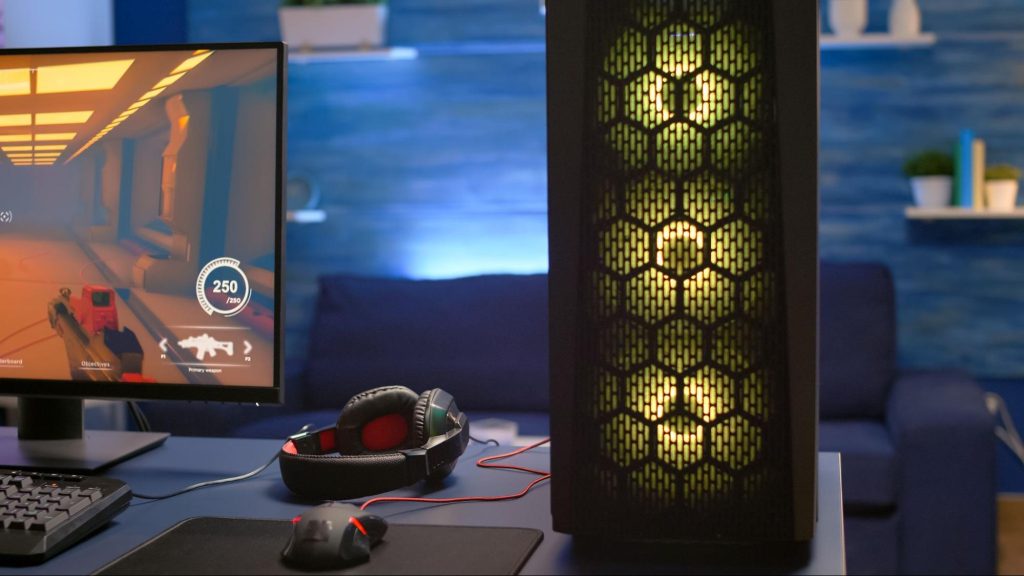 Imagem de uma fotografia colorida em que há um quarto, com paredes azuis ao fundo, e no primeiro plano uma mesa cinza com um CPU com luzes amarelas e, do outro lado, um fone de ouvido, um mouse, ambos pretos e vermelhos, e com um monitor reproduzindo um jogo na tela.