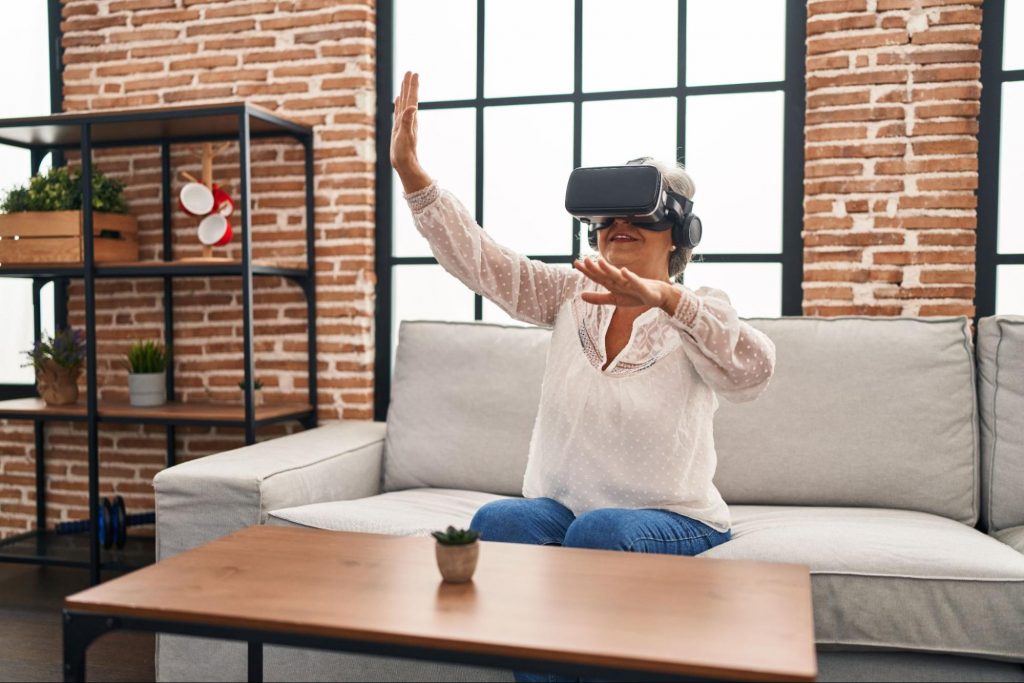 Mulher idosa sentada em sofá com um óculos de jogos de realidade virtual, fazendo movimentos com os braços