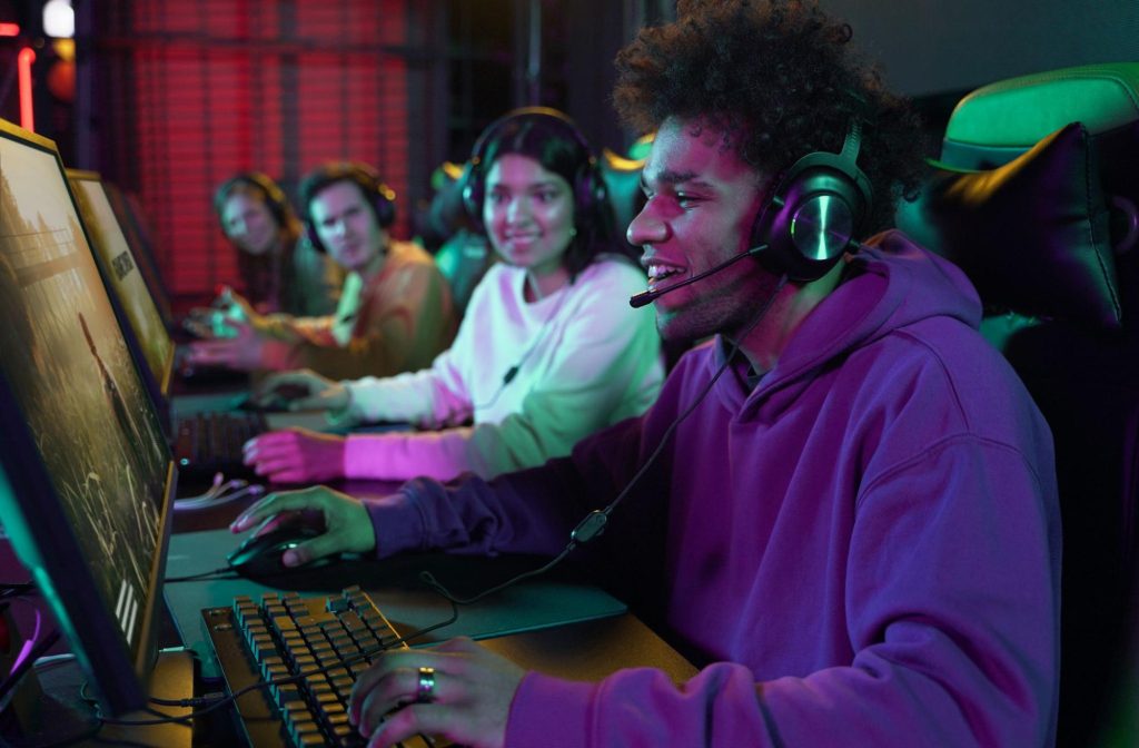Imagem de uma sala de jogos com vários computadores gamers e com quatro jovens jogando em cada um, em primeiro plano, tem um homem negro com fones de ouvido jogando em um computador gamer.
