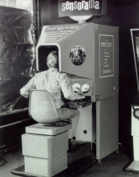 Imagem de uma fotografia em preto e branco em que um homem está sentado em uma cadeira de frente para uma máquina enorme que parece uma cabine de fotos, o Sensorama.