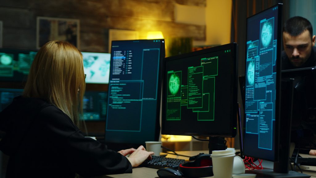 Imagem de uma mulher loira de costas mexendo em um computador, há três telas em frente dela e todas apresentam códigos de cibersegurança. A imagem representa a tentativa de proteção contra os tipos de ransomware.