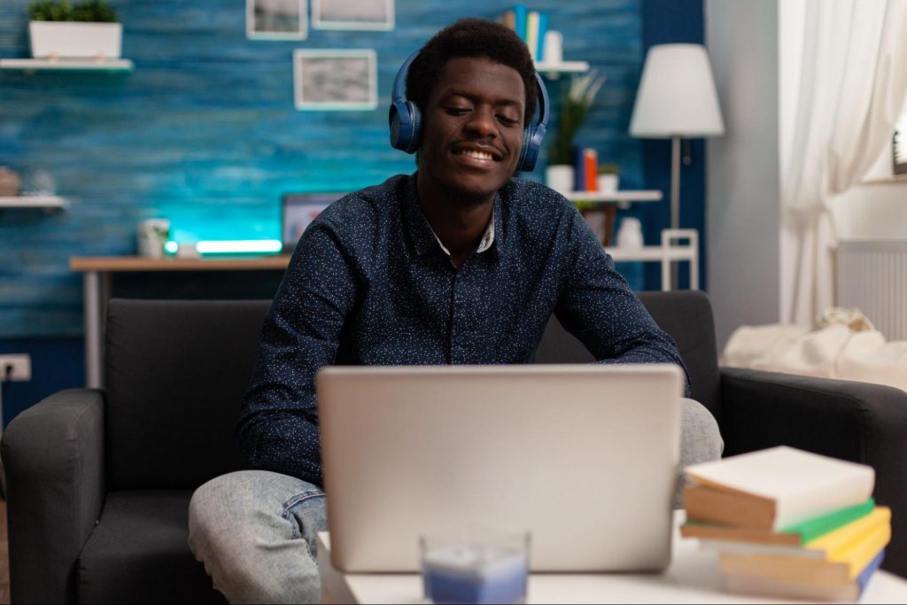 Imagem de uma fotografia em que há um homem negro sentado em um sofá, dentro de uma sala e de frente para um notebook, a imagem simula o modelo de trabalho home office.