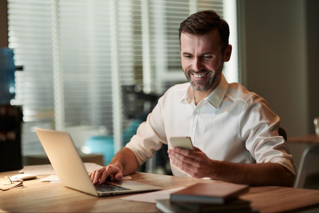 Imagem de uma fotografia em que há um homem branco sentado de frente para um notebook, sorrindo, teclando nele com uma mão e com a outra verificando o celular. 