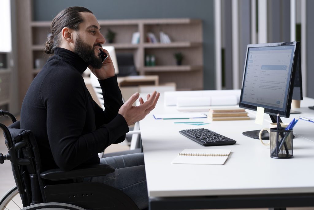 Profissional de TI, utilizando cadeira de rodas, enquanto conversa ao celular, com computador, cadernos e canetas a sua frente.