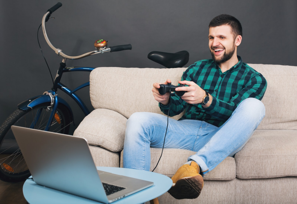 um homem branco com barba e cabelo castanhos está sentado sobre um sofá creme, vestindo uma calça jeans, botas marrom e uma camisa quadriculada entre preto e verde. ele segura o console de um videogame e olha para a tela de um notebook, que está aberto em sua frente, sobre uma mesa baixa de centro. Ao fundo, uma parede preta e uma bicicleta são mostradas