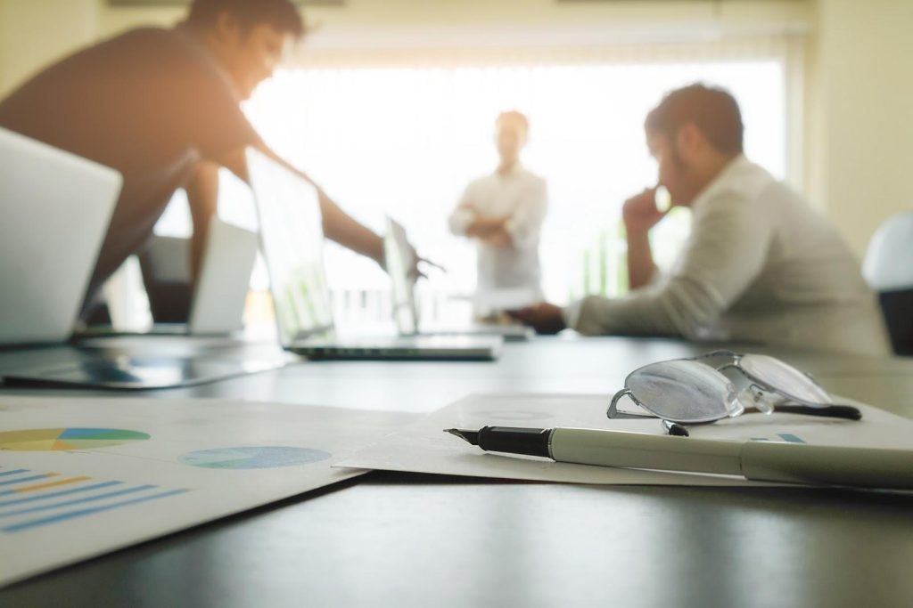 a mesa de um escritório é mostrada em foco. Sobre ela, papéis e canetas estão dispostos e, ao fundo, três pessoas com roupas sociais estão em pé ao redor da mesa, discutindo ideias com 3 desktops abertos