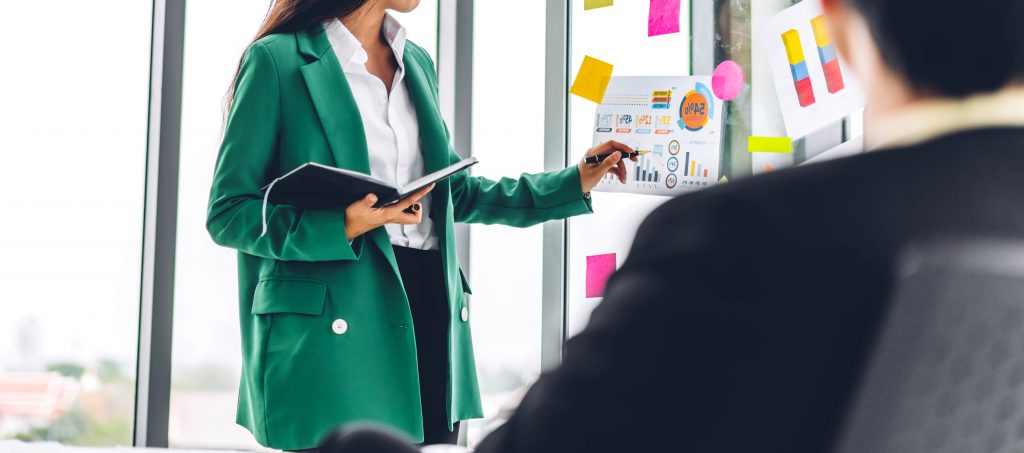 mulher aponta para um quadro com gráficos e post-its enquanto segura um caderno na outra mãos