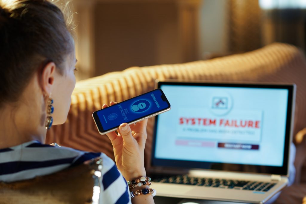 Mulher com celular na mão, falando com um chatbot. Ao fundo, um notebook com “system failure” na tela
