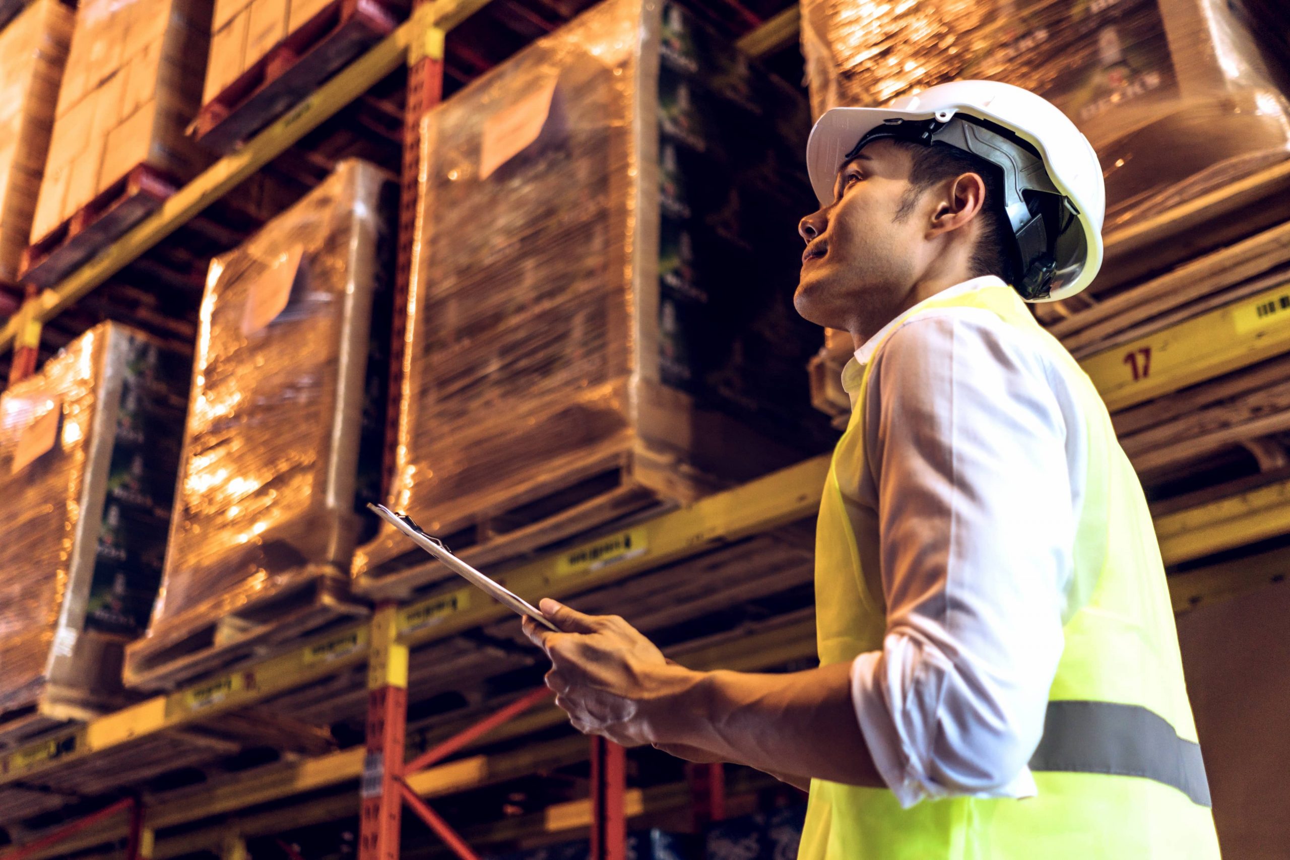 o que é supply chain capa Homem asiático com EPIs e prancheta em mãos olhando para cima dentro de um depósito