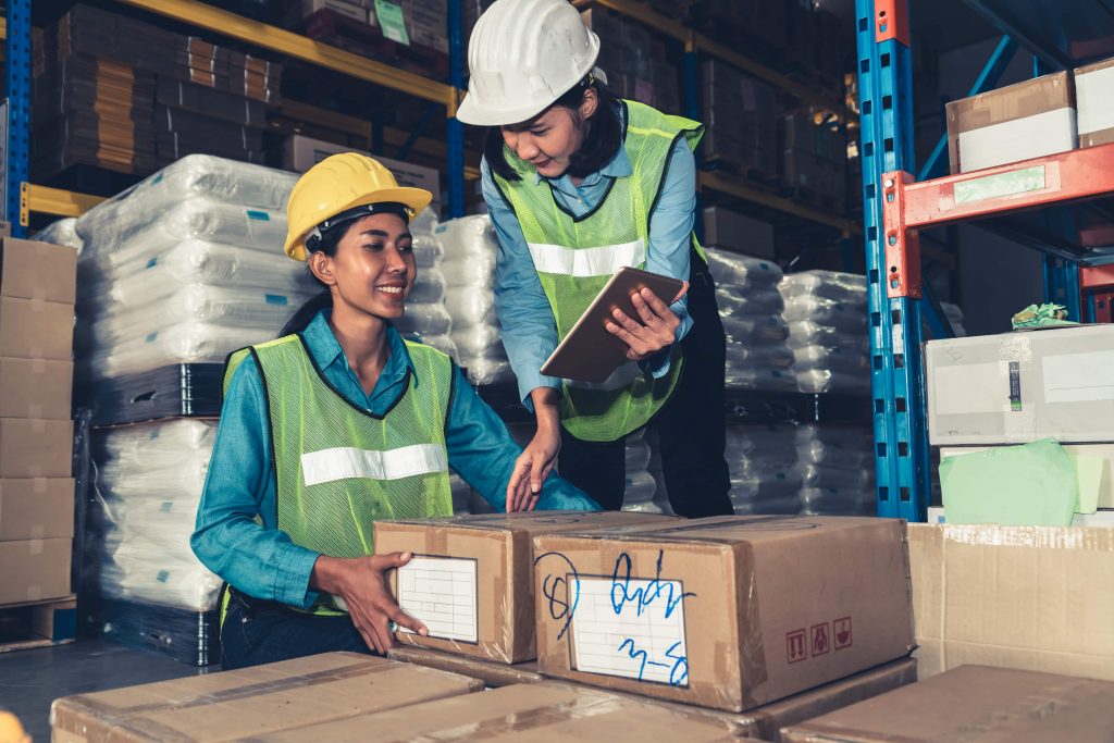 Duas mulheres asiáticas com EPIs trabalham em um estoque. Uma delas possui um tablet em mãos, a outra segura uma caixa. 