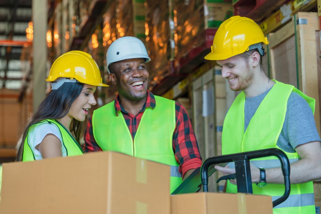 Mulher branca, homem negro e homem branco usando EPIs com prancheta em mãos para gerenciar as caixas de armazém à frente.