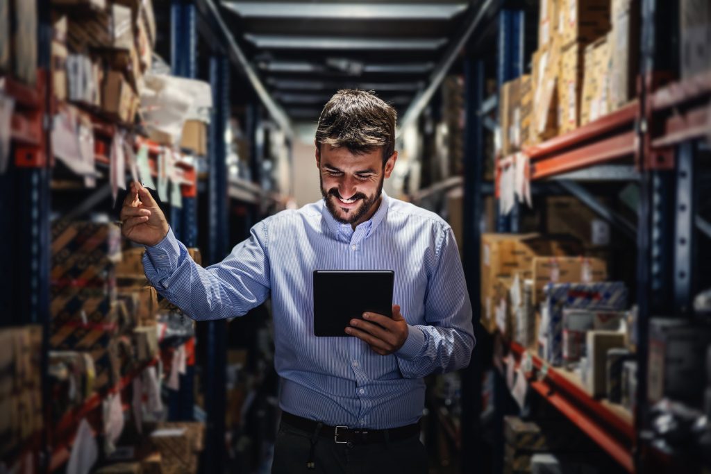 Homem branco com tablet na mão verifica as prateleiras de um estoque.