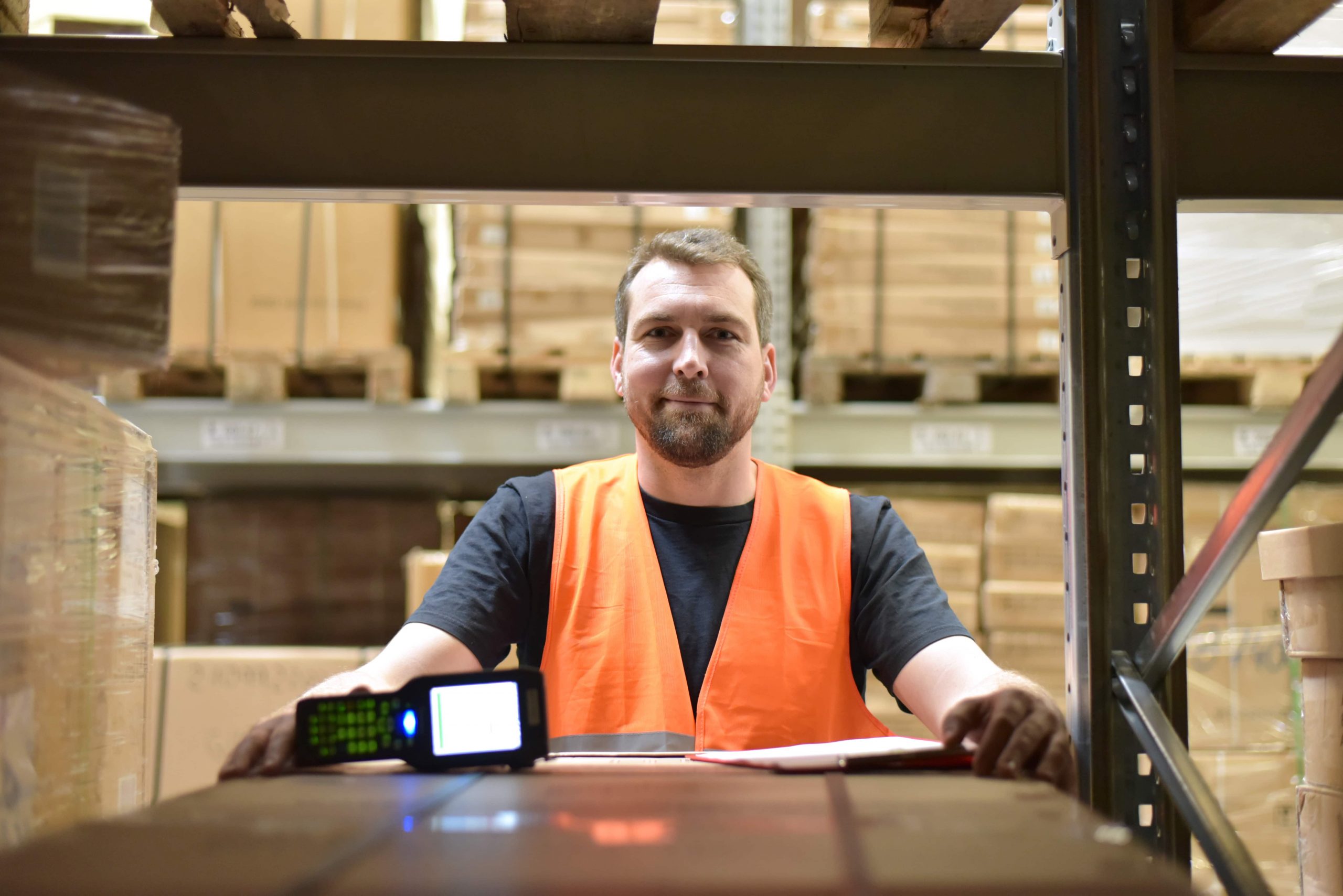 Sistema WMS capa Homem branco de colete entre caixas de um armazém, com uma prancheta e um leitor de código à sua frente.