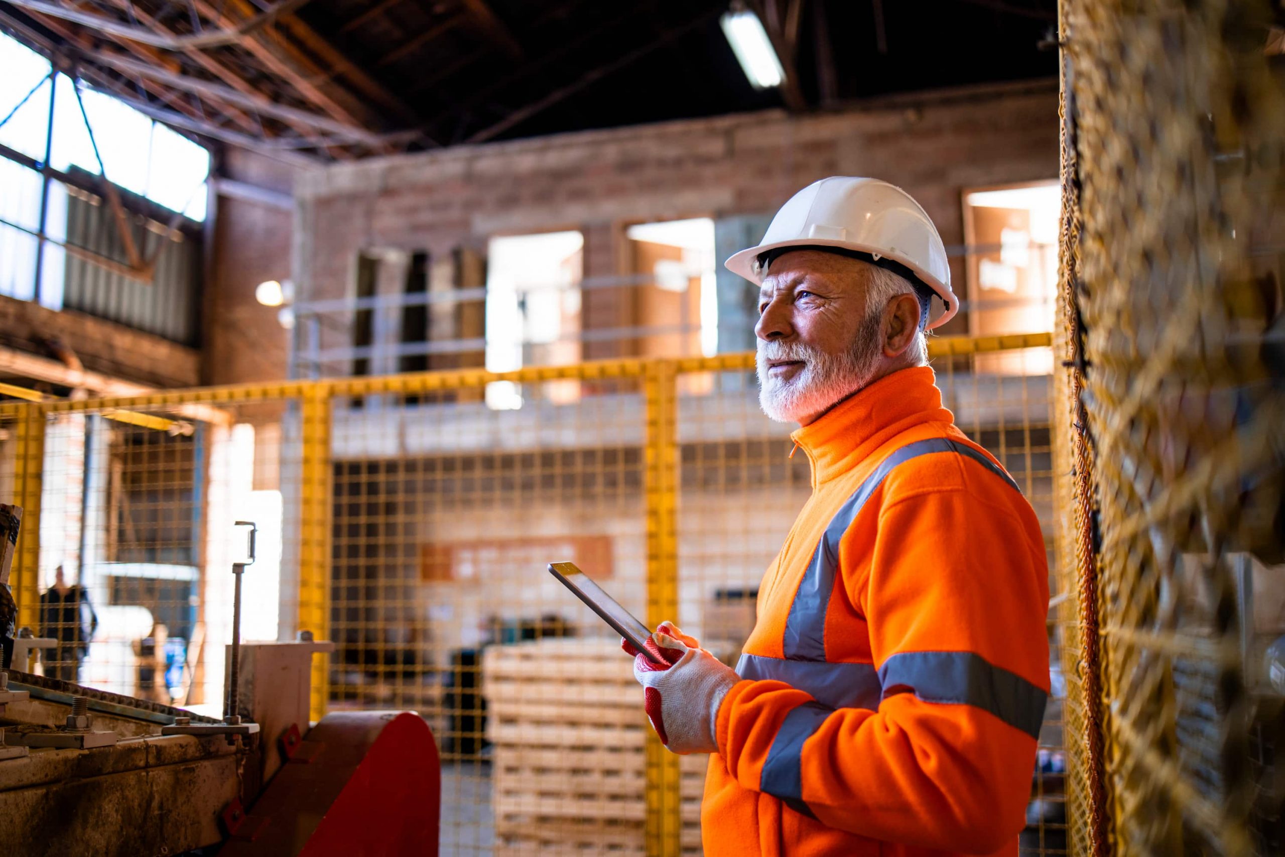 indústria 5.0 capa Homem branco grisalho com EPIs segurando um tablet dentro de uma fábrica.