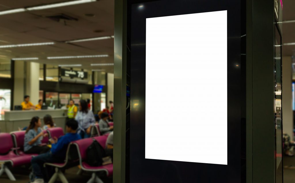 Tela em branco à frente e, ao fundo, diversas cadeiras de espera com pessoas em desfoque sentadas. 