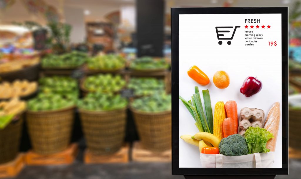 Telão em um mercado apresenta imagens de frutas para compra. Ao fundo, em desfoque, diversas cestas de vegetais estão dispostas. 