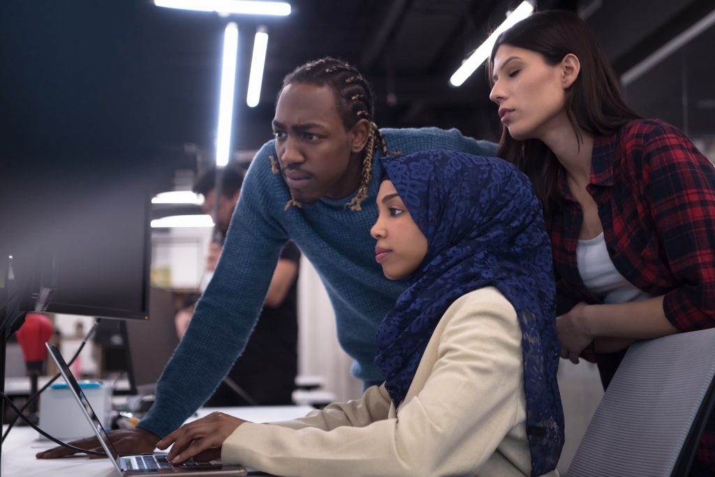 Mulher negra de lenço, homem negro e mulher branca em um escritório olhando juntos para a tela de um notebook.
