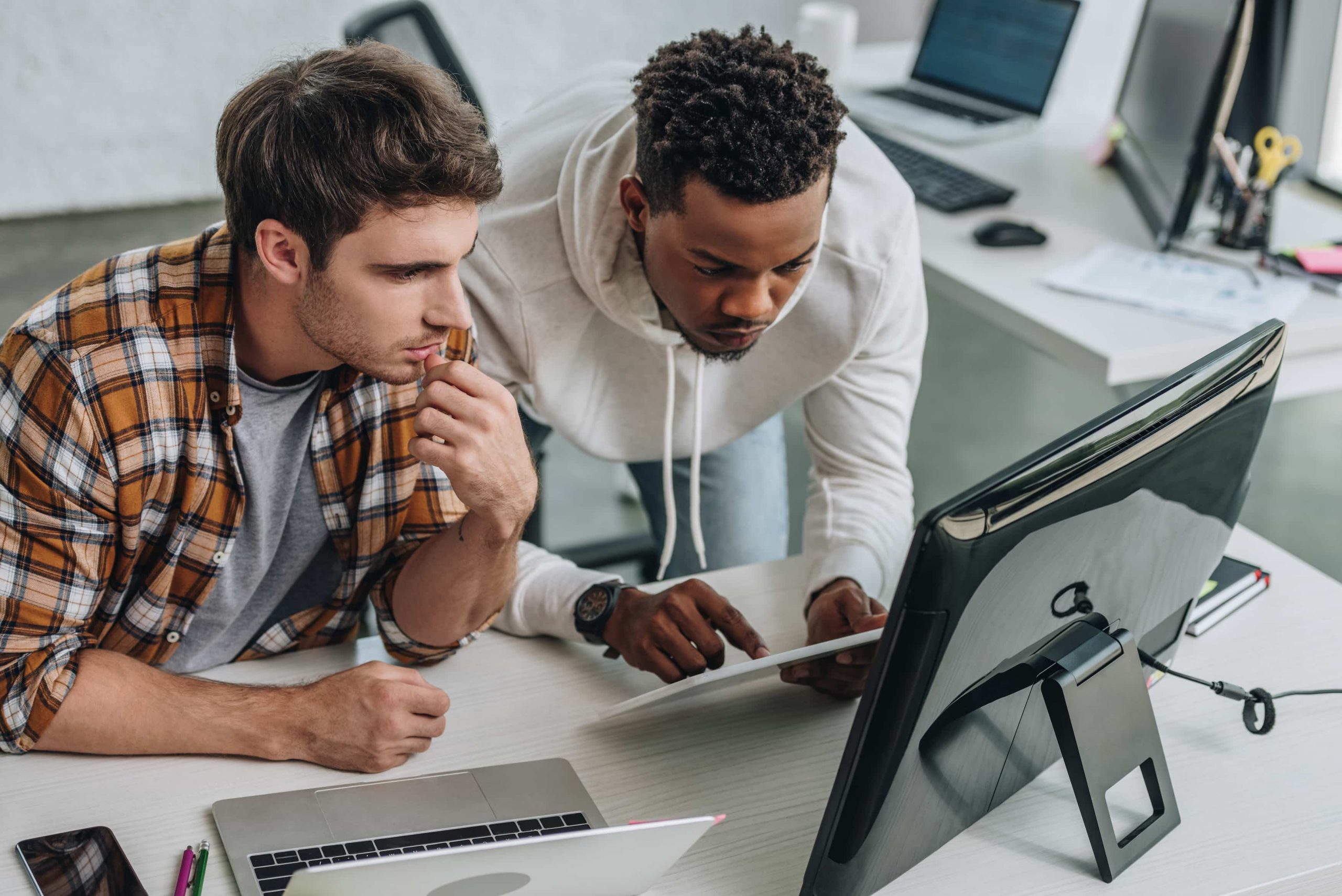 Homem negro e homem branco sentados, encarando preocupados a tela de um computador.