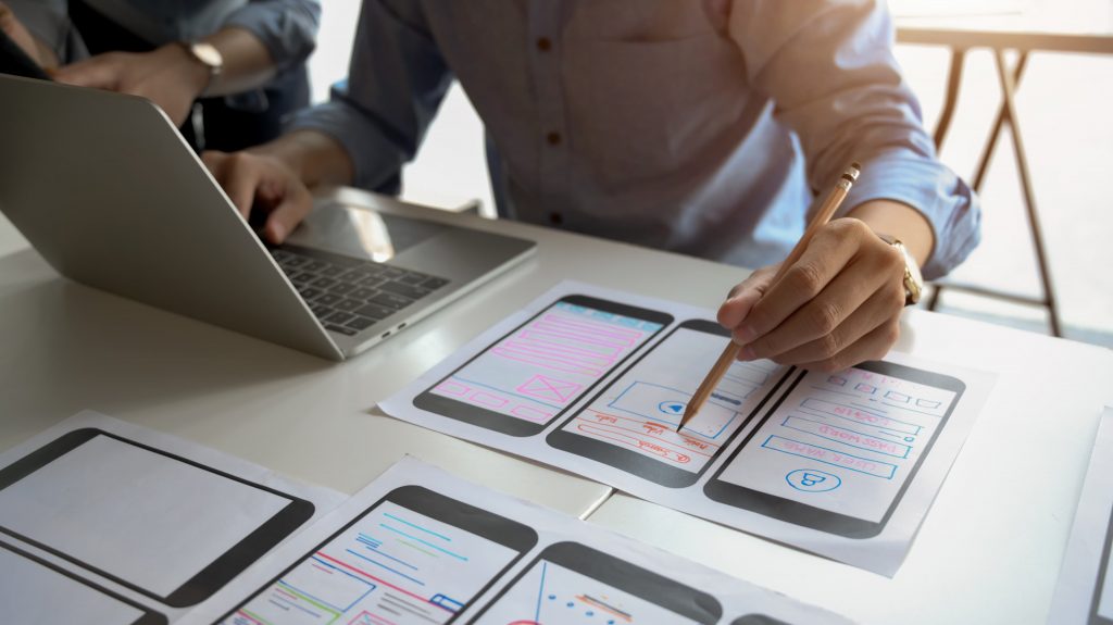 Pessoa com notebook à frente e lápis em mão, organizando papéis que contém desenhos de design de apps