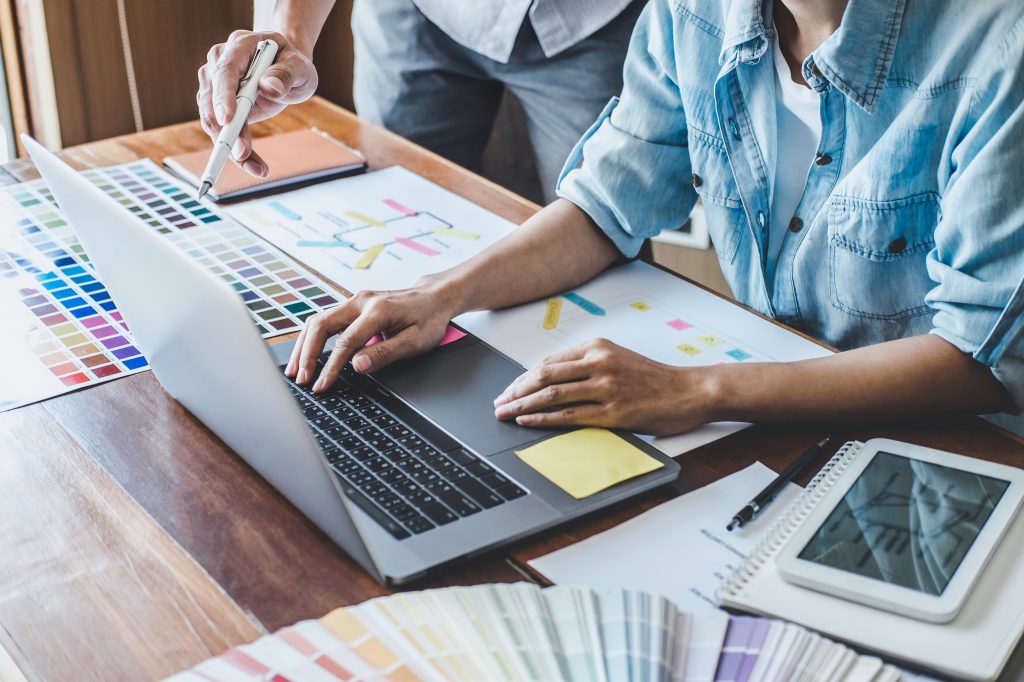 Duas pessoas com um notebook à frente, junto a paleta de cores e demais folhas de desenvolvimento de site a partir de front end.