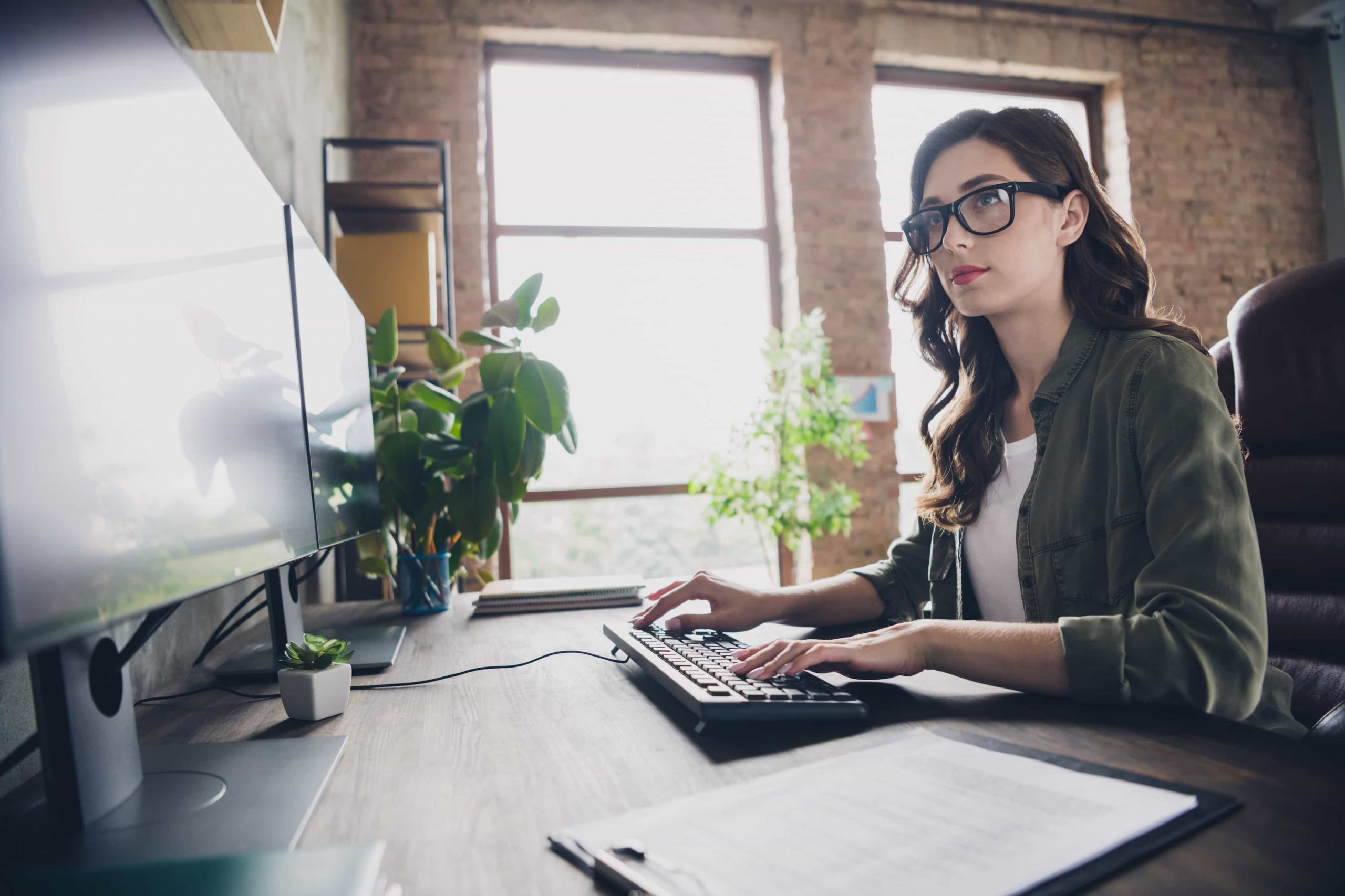 Mulher branca de óculos digita em um teclado e olha para os monitores à frente enquanto programa.