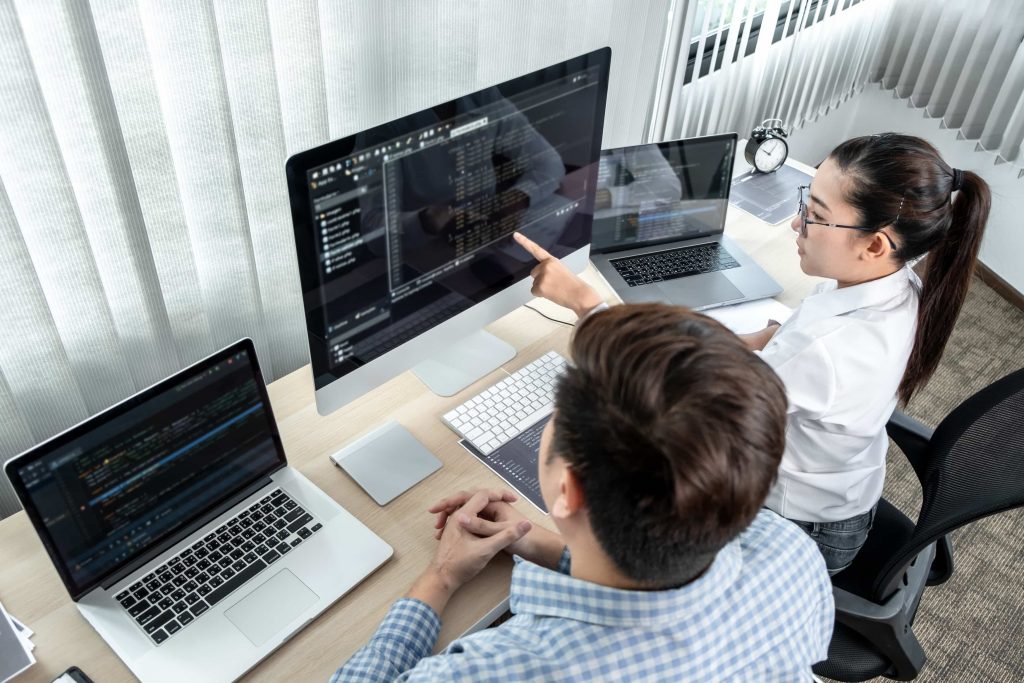 Homem e mulher sentados frente a três computadores, analisando códigos para cibersegurança.
