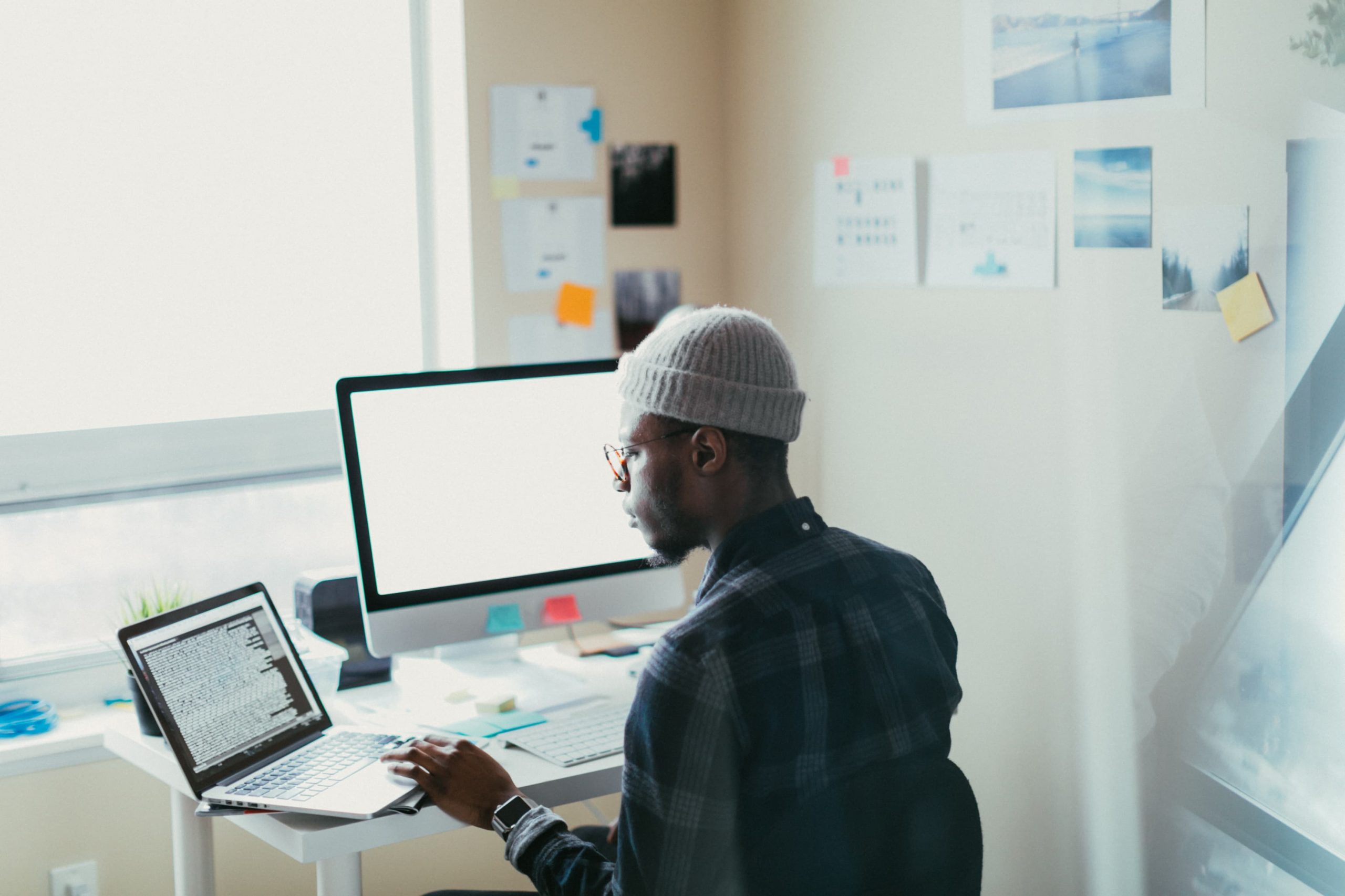 O que é back end capa homem negro de touca e óculos de costas, sentado em uma escrivaninha com notebook e monitor à frente.